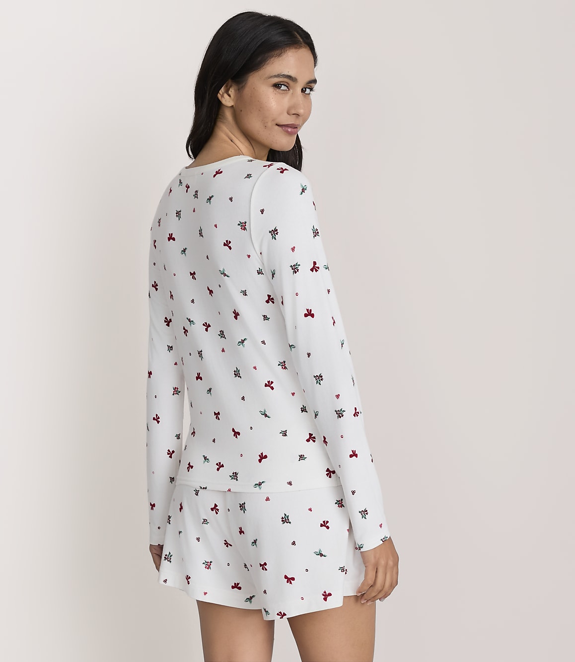 A woman wearing white pajamas with a small red and green pattern stands against a plain, light-colored background.