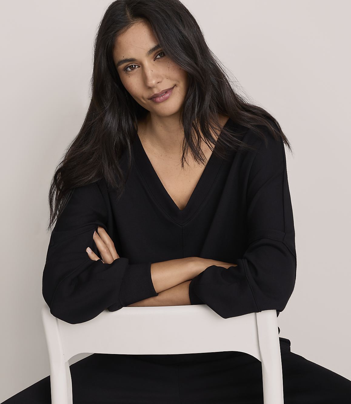 A woman with long dark hair wearing a black V-neck top leans on a white chair against a plain light-colored background.