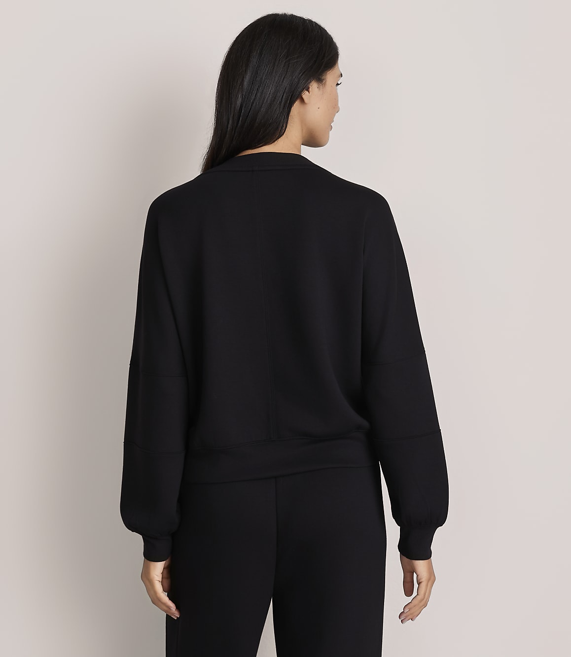 A woman with long dark hair stands with her back to the camera wearing a black long-sleeved top and black pants against a plain light-colored background.