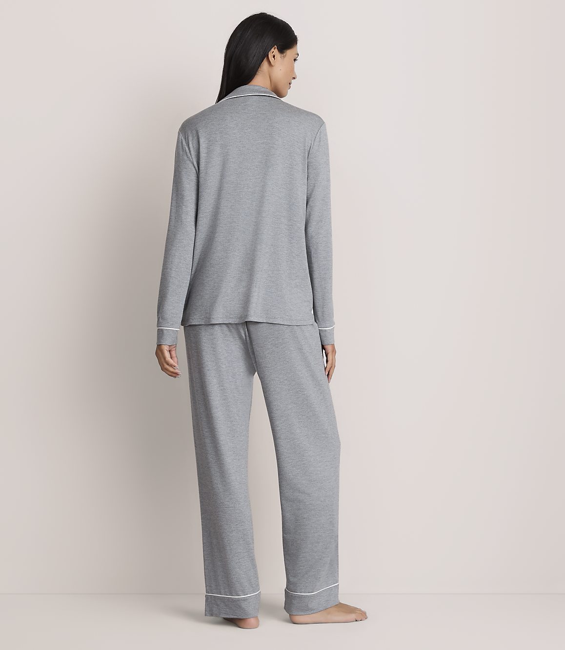A woman stands with her back to the camera wearing grey pajamas against a plain, light-colored wall.