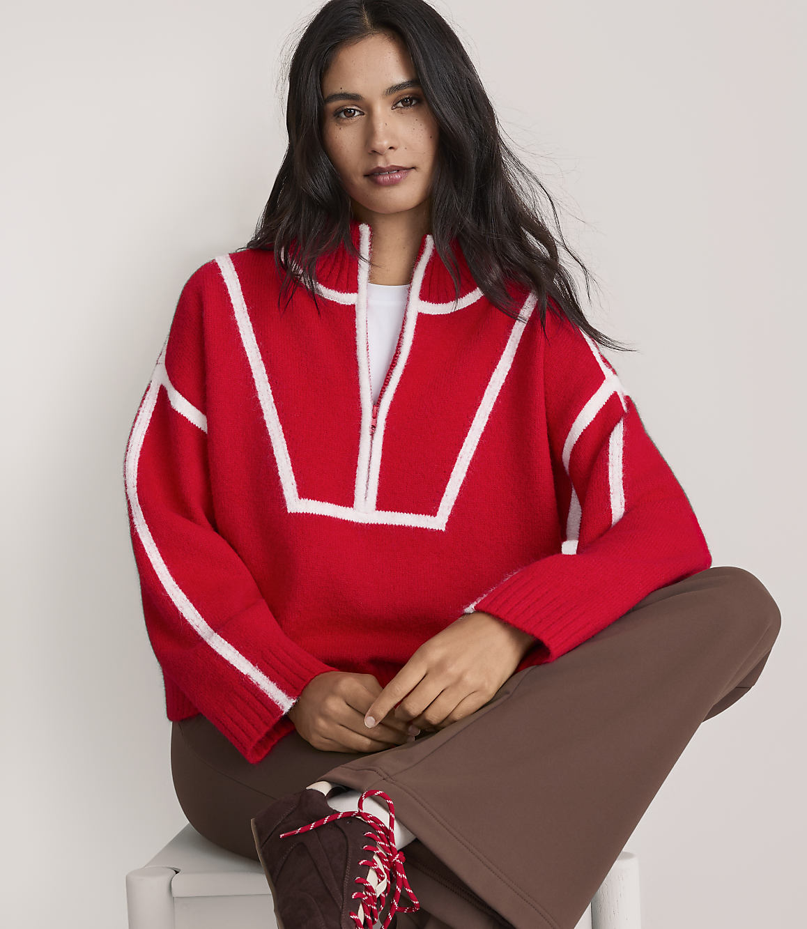 A woman wearing a red and white patterned sweater and brown pants sits against a plain white background.