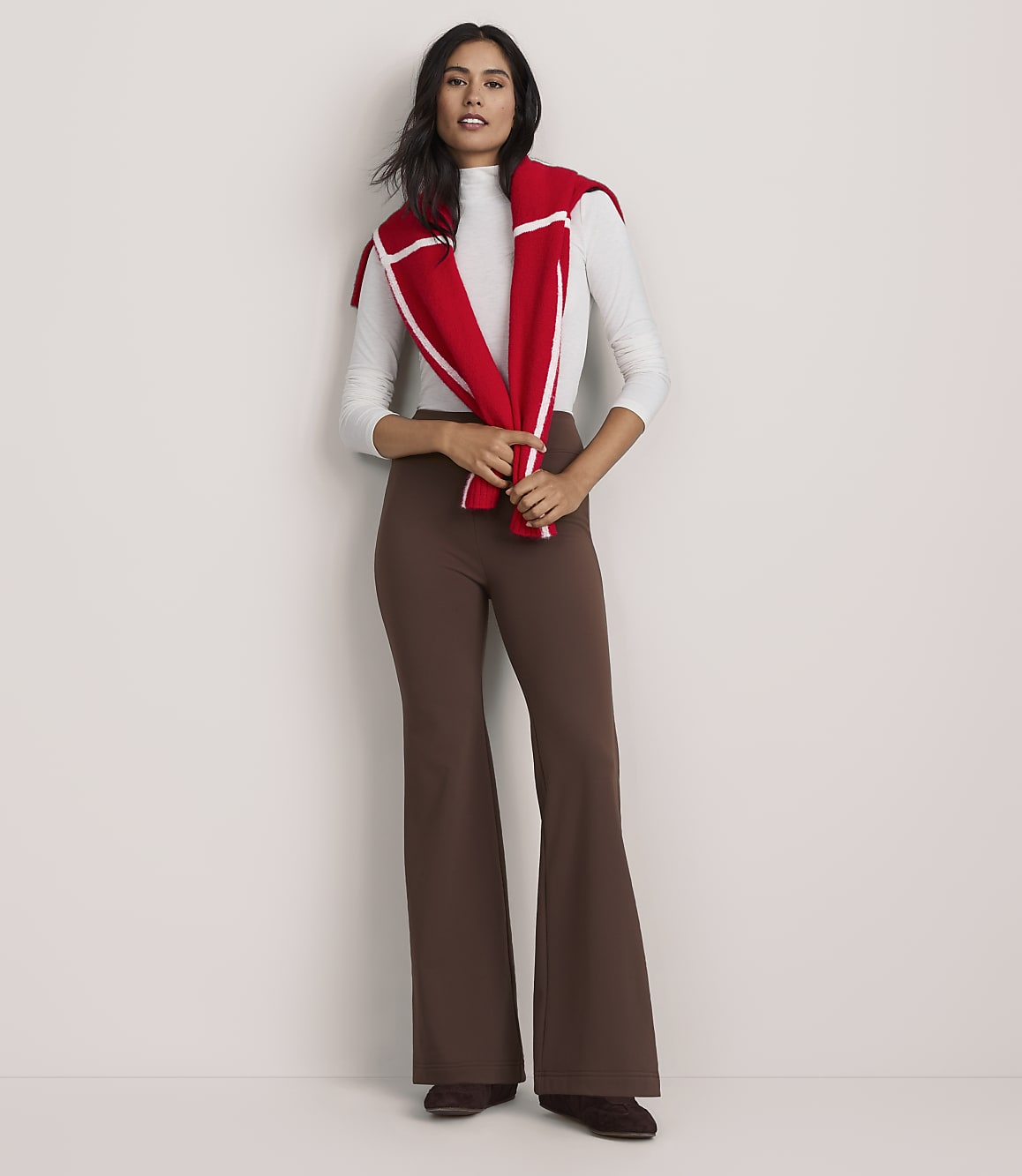 A woman wearing a white top, brown flared trousers, and a red sweater draped over her shoulders stands against a plain white background.