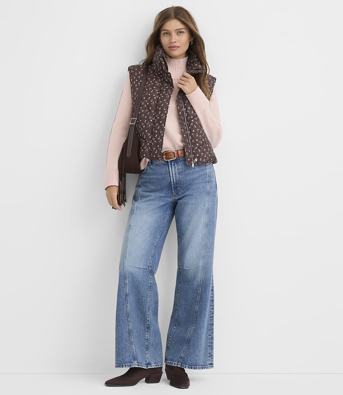 A woman wearing a brown patterned vest, pink sweater, and flared blue jeans stands against a plain white background.
