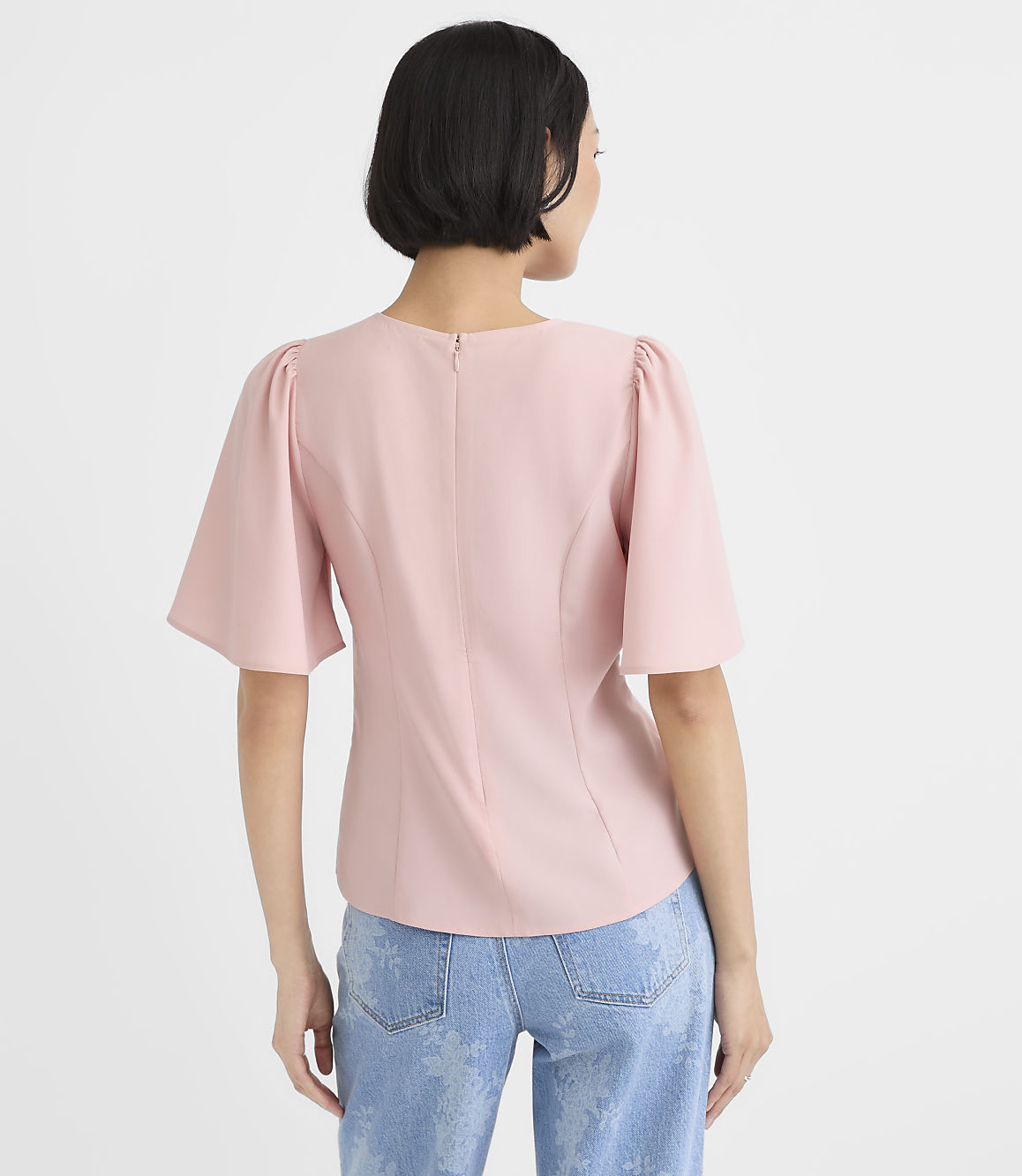A woman wearing a light pink puff-sleeve top and blue jeans stands with her back to the camera against a plain white background.