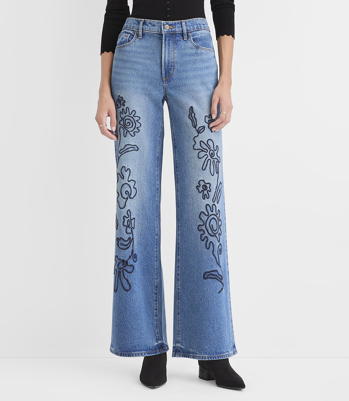 A person wears blue wide-leg jeans with black floral embroidery against a plain white background.