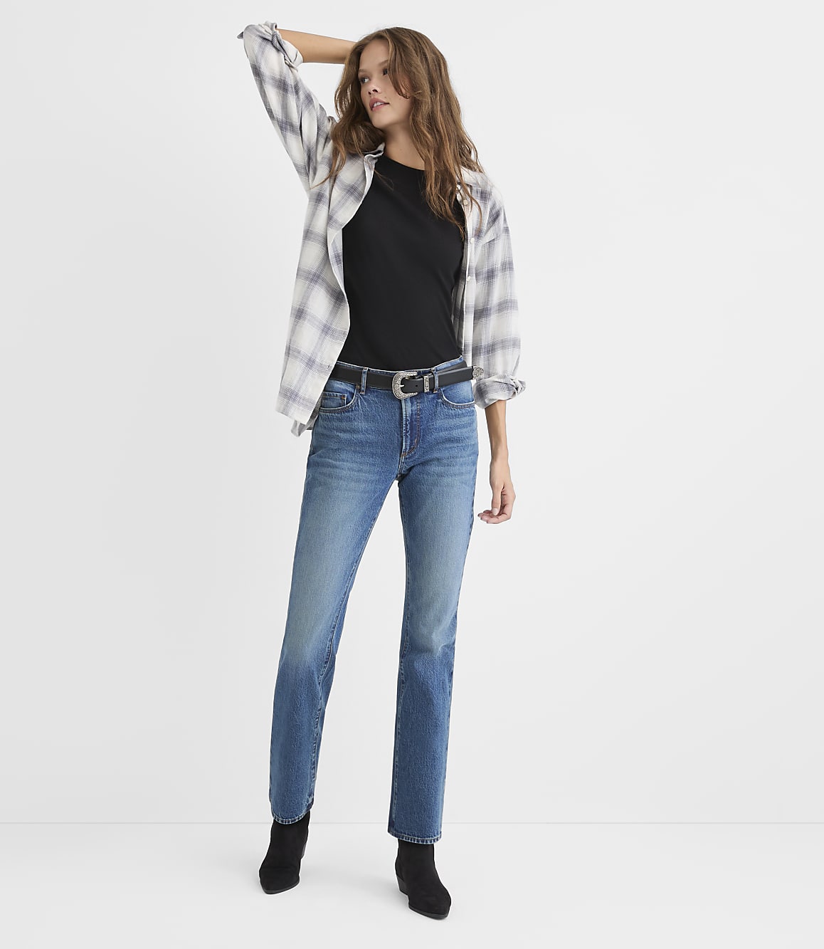 A woman wearing a black t-shirt, an open plaid shirt, blue jeans, and black boots stands against a plain white background.
