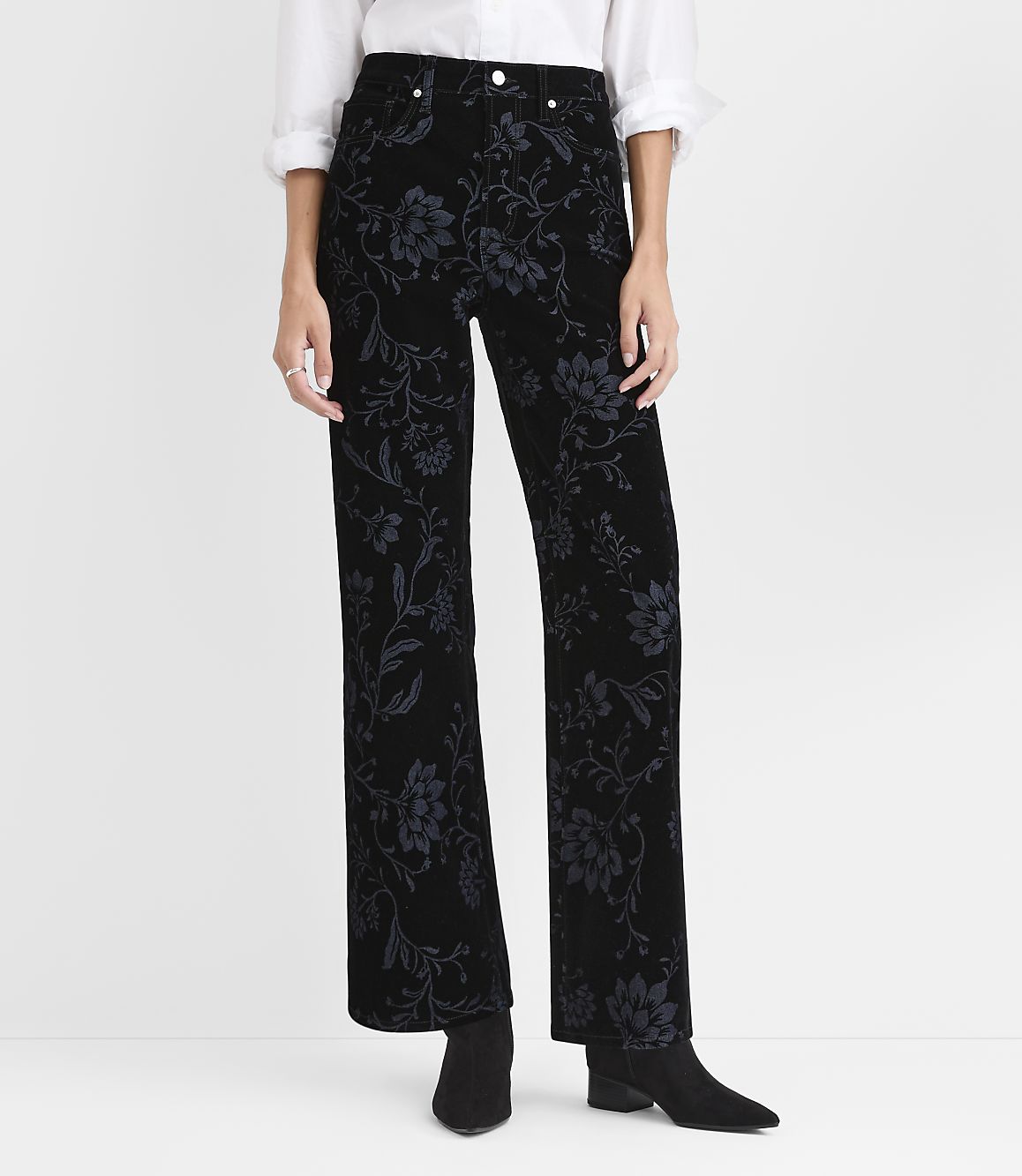 A person wears black floral velvet trousers, a white shirt, and black boots against a plain white background.