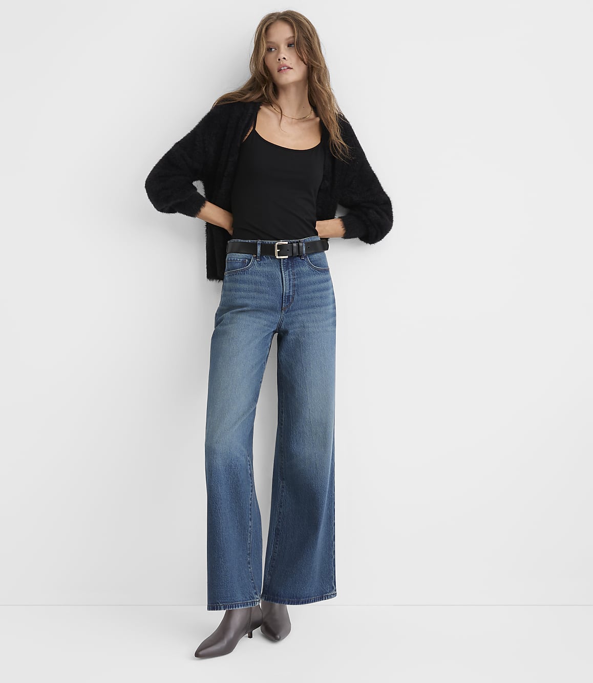 A woman wearing a black cardigan, black top, and wide-leg blue jeans stands against a plain white background.