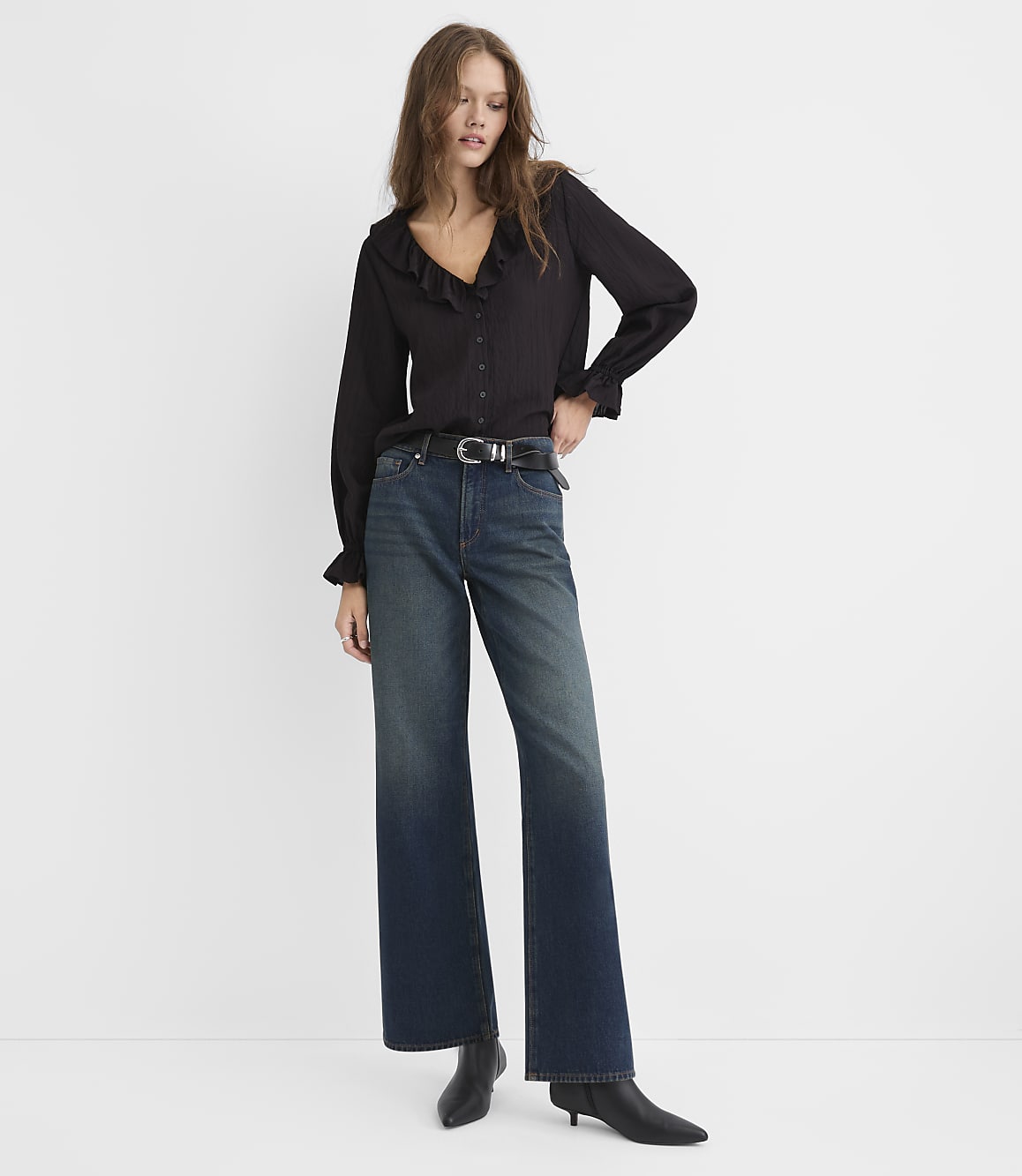 A woman wearing a black ruffled blouse and wide-leg blue jeans stands against a plain white background.