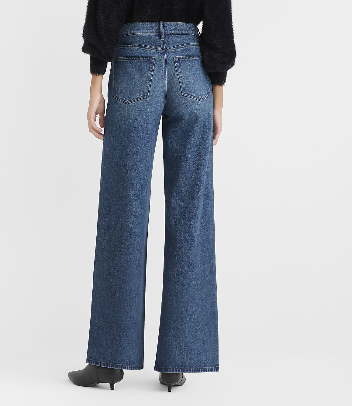A person wearing a black fuzzy sweater, wide-leg blue jeans, and black pointed heels stands against a plain white background.