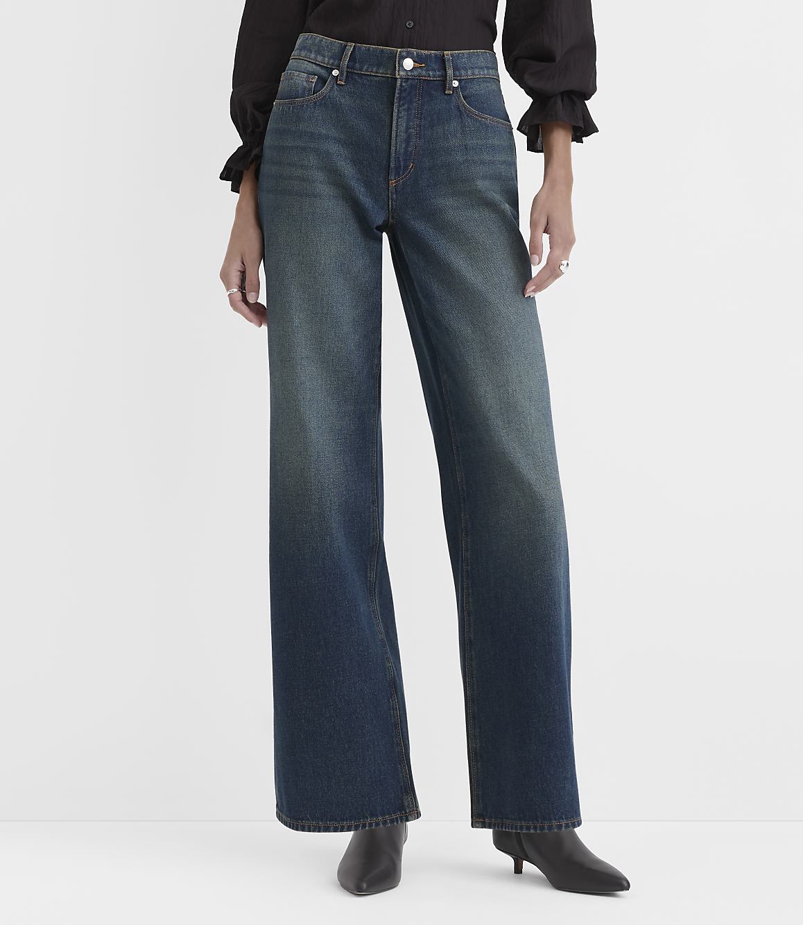 A person wearing a black blouse, wide-leg blue jeans, and black pointed-toe boots stands against a plain white background.