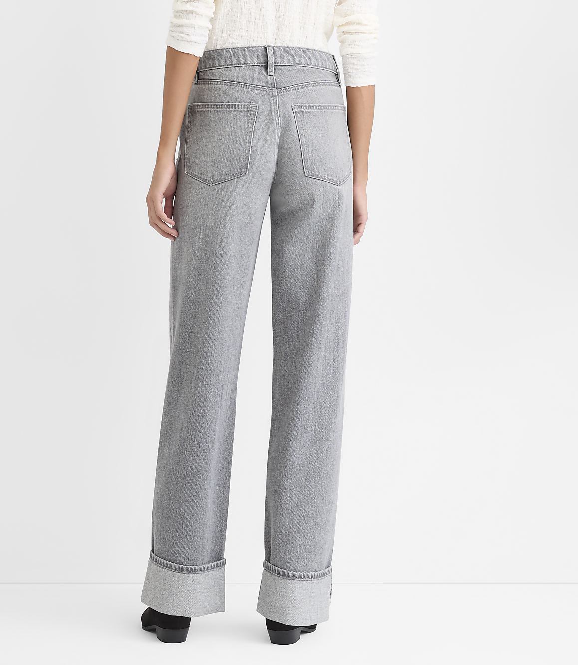 A person stands facing away from the camera wearing wide-leg grey jeans and a white top against a plain white background.