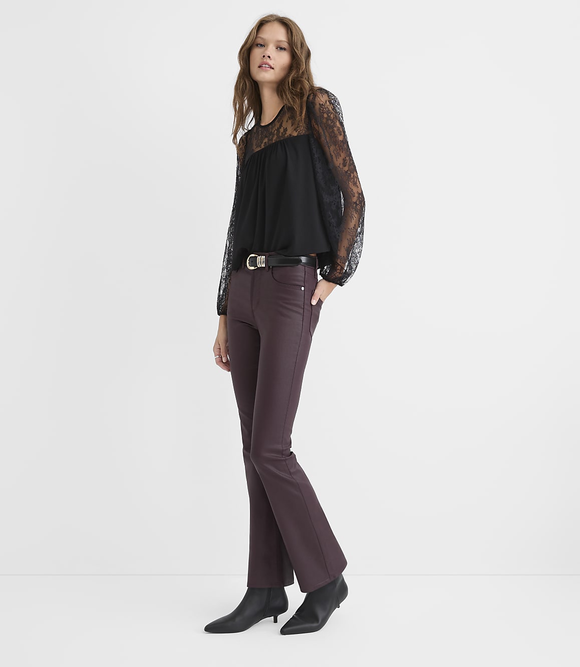 A woman wearing a black lace-sleeved top and burgundy flared trousers stands against a plain white background.