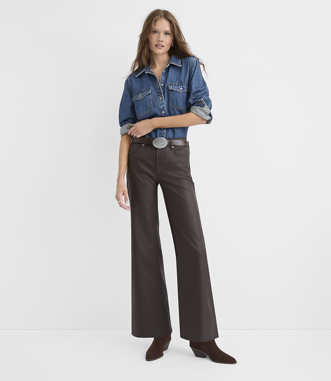 A woman wearing a denim shirt, brown wide-leg trousers, and brown boots stands against a plain white background.