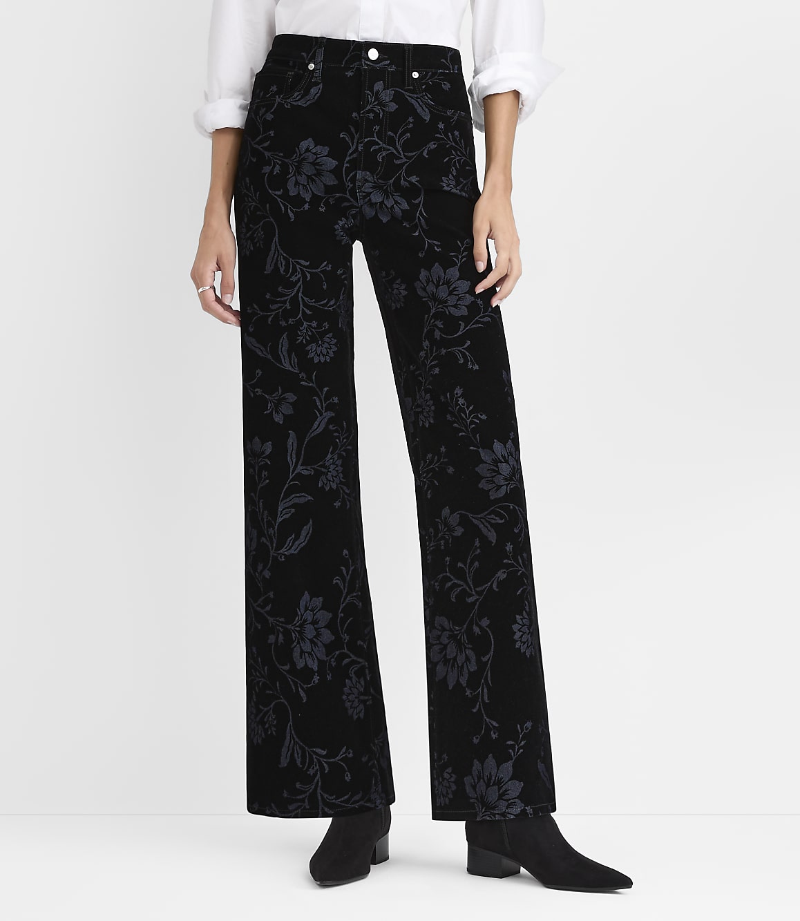 A person wearing a white shirt and black floral-patterned wide-leg pants stands against a plain white background.