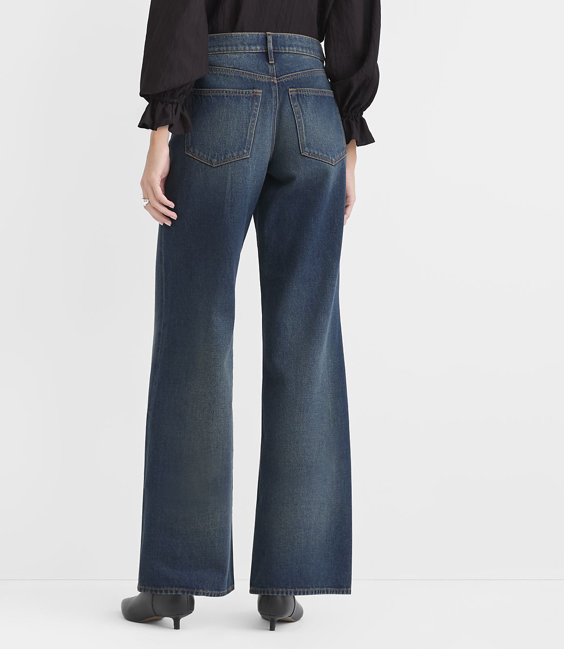 A person wearing a black top and wide-leg blue jeans stands against a plain white background.