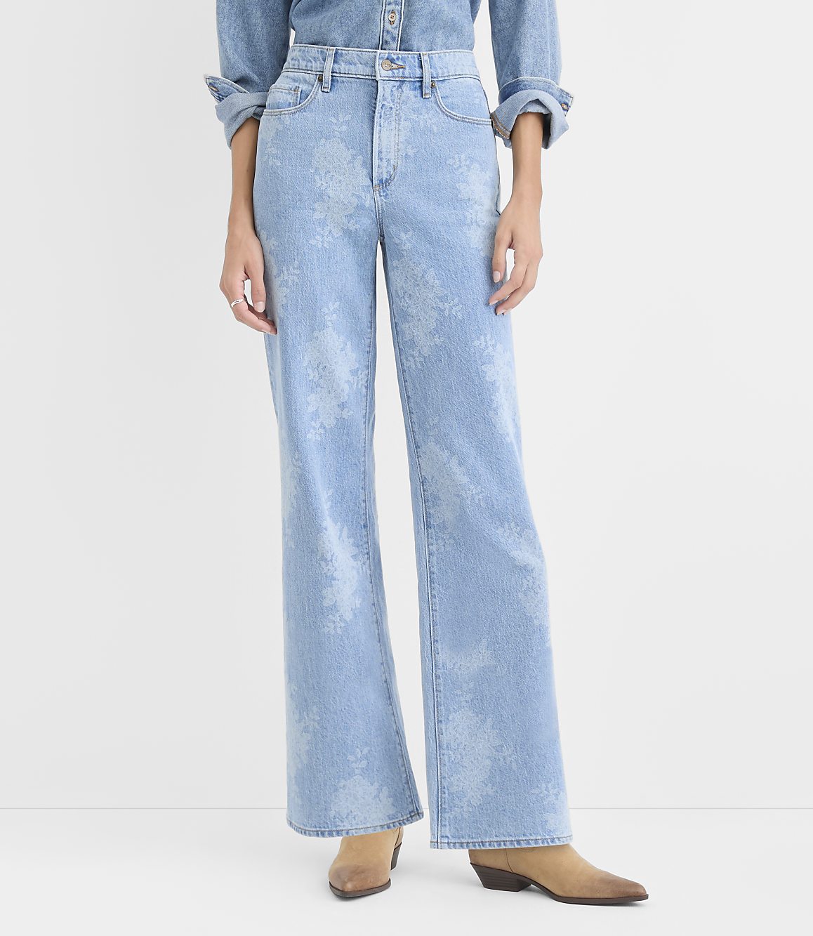 A person wearing a light-wash denim shirt and floral-patterned wide-leg jeans stands against a plain white background.