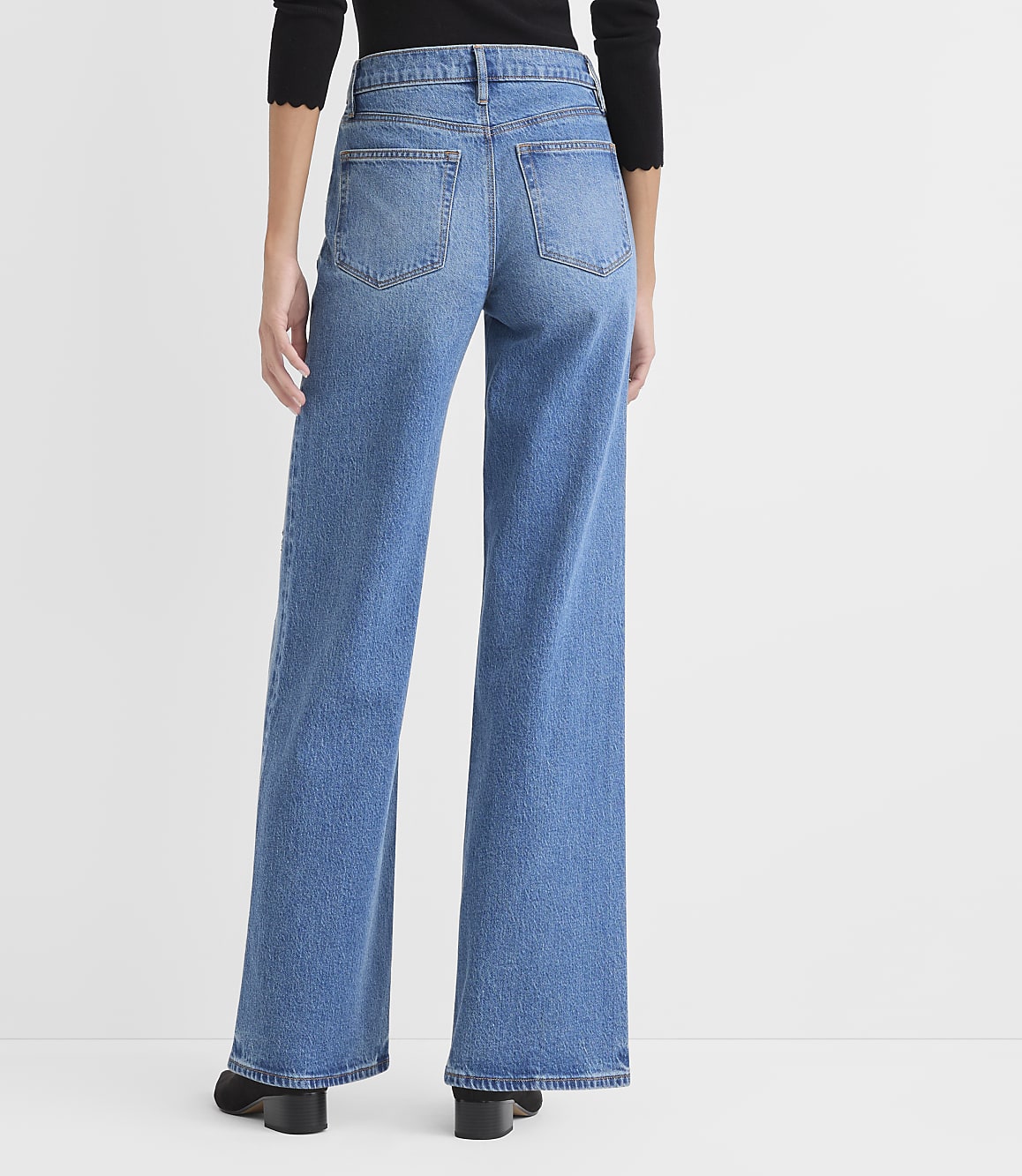 A person wearing a black top and wide-leg blue jeans stands against a plain white background.