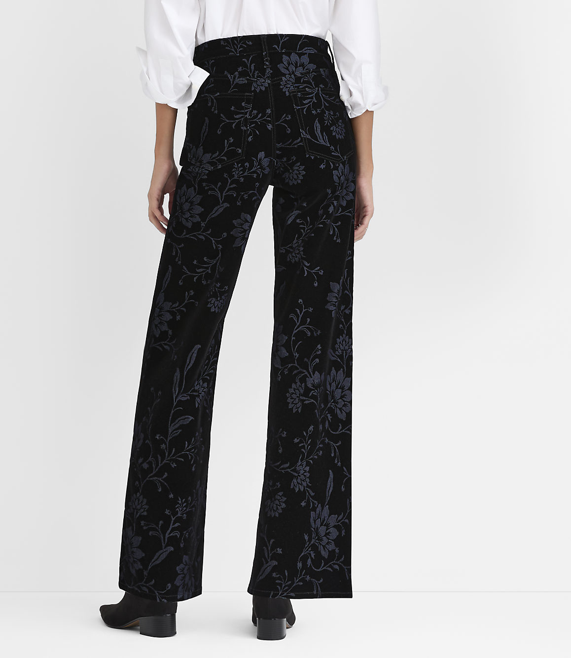 A person stands with their back to the camera wearing a white blouse and floral-patterned black velvet trousers against a plain white background.