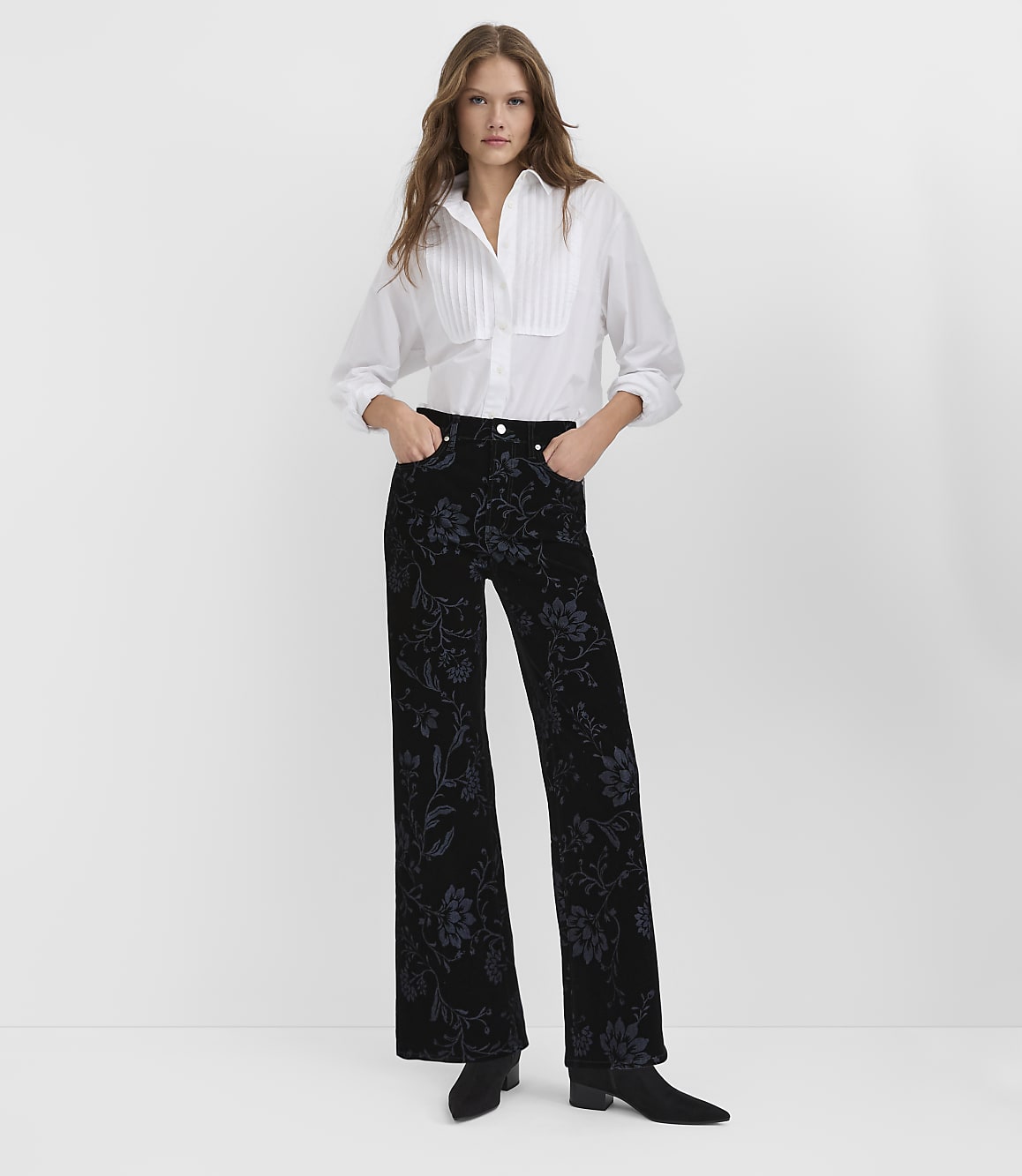 A woman wearing a white pleated blouse and floral black velvet trousers stands against a plain white background.