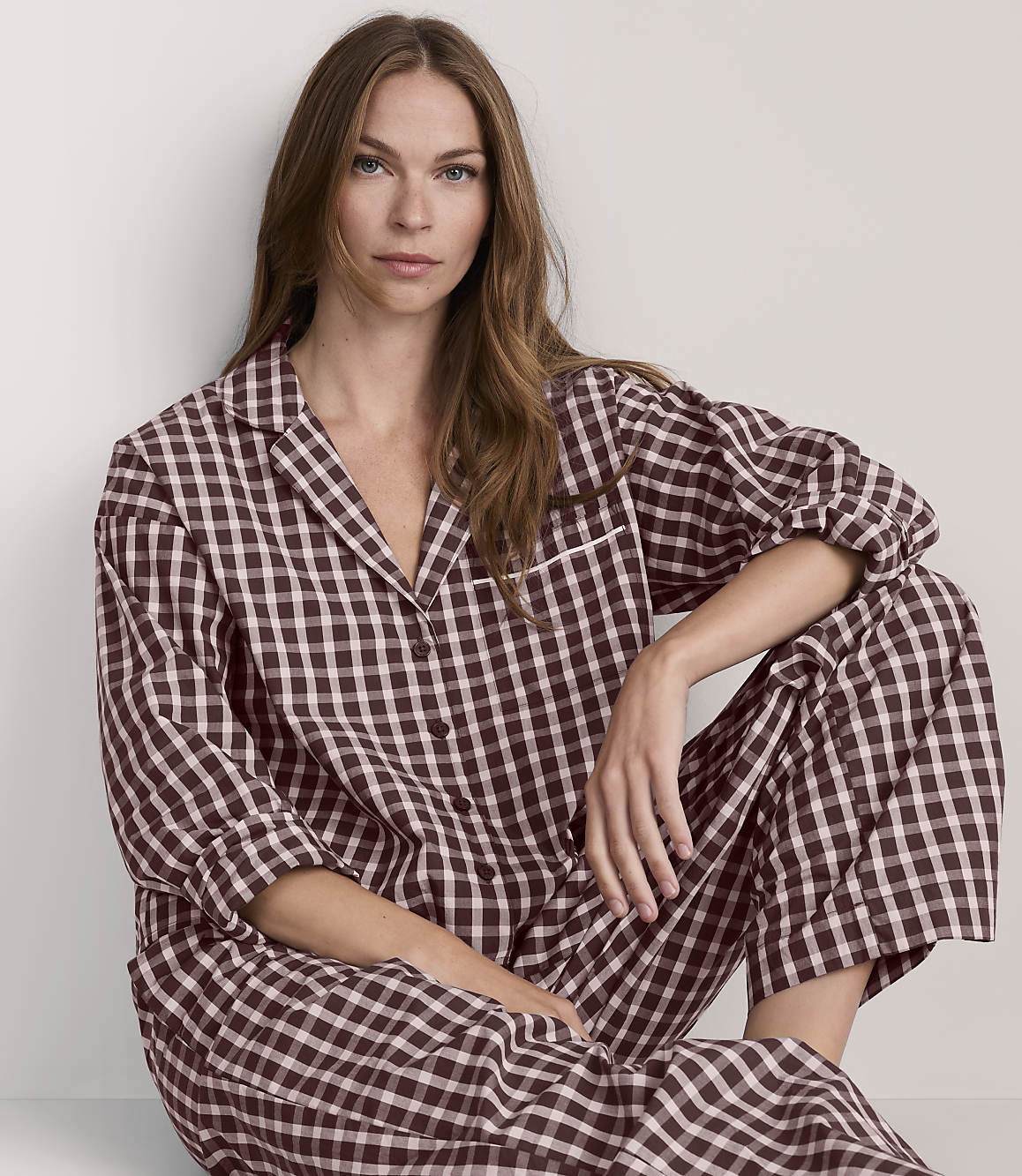 A woman in brown and white checkered pajamas sits against a plain, light-colored background.