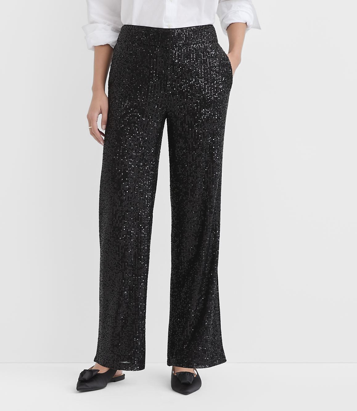 A person wearing a white shirt, shimmering black sequined trousers, and black pointed flats stands against a plain white background.