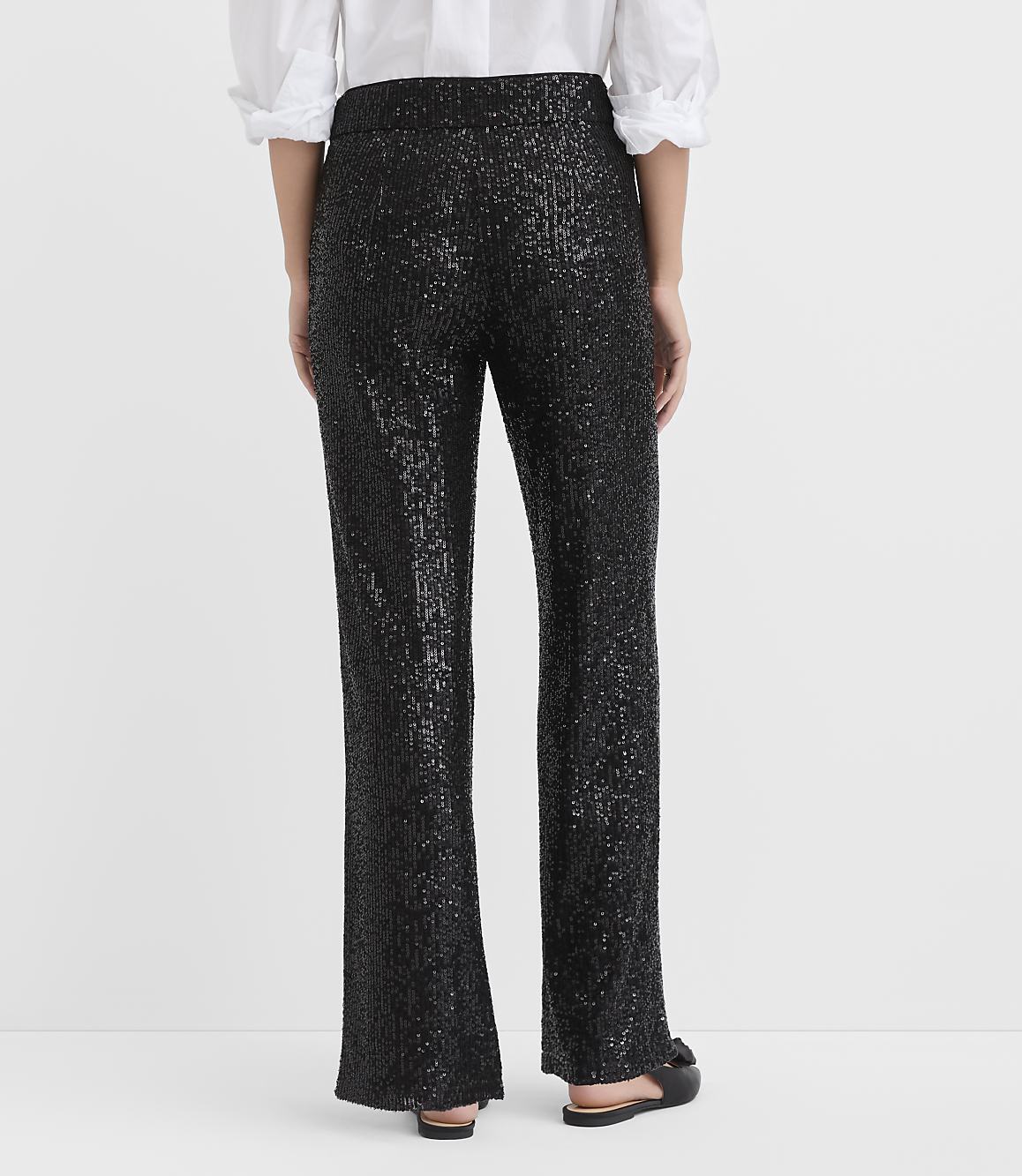 A person stands with their back to the camera wearing a white blouse and black sequined trousers against a plain white background.