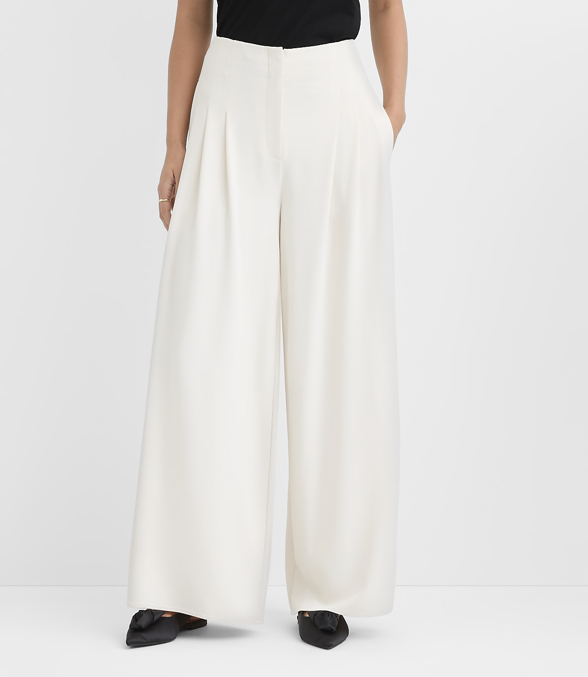 A person wearing a black top and wide-leg white trousers stands against a plain white background.