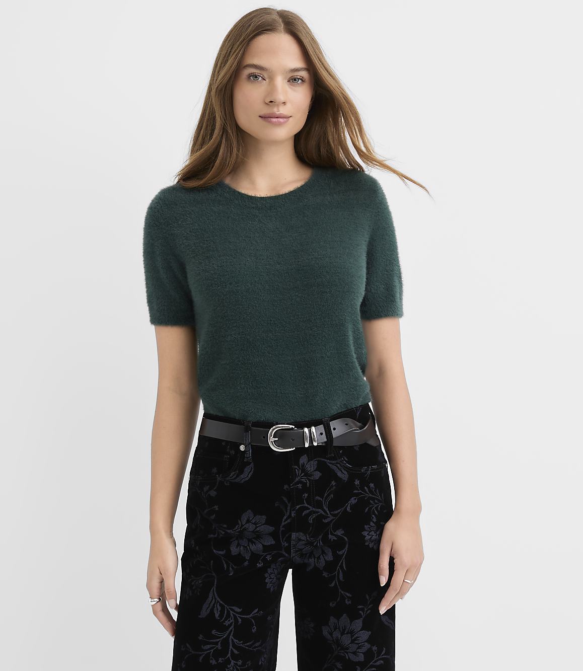 A woman wearing a dark green fuzzy short-sleeved sweater and floral black pants stands against a plain white background.