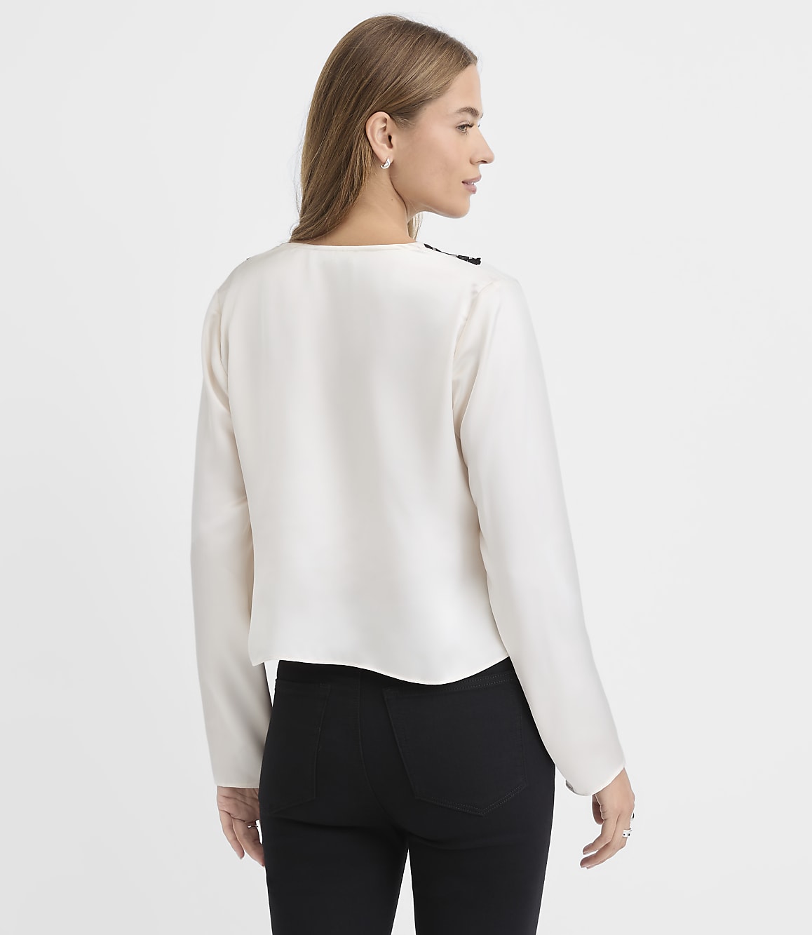 A woman wearing a white long-sleeved blouse and black pants stands with her back to the viewer against a plain white background.