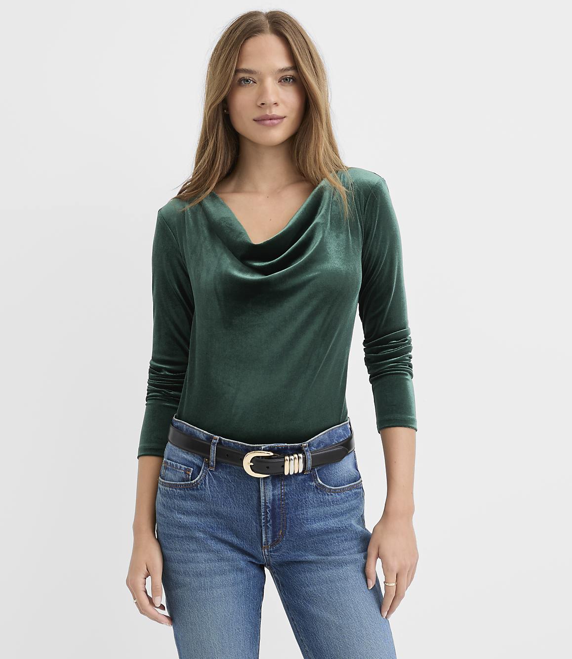 A woman wearing a dark green velvet cowl-neck top and blue jeans stands against a plain white background.