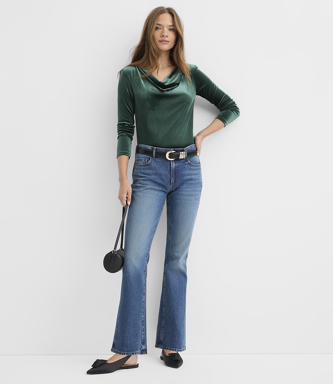 A woman wearing a green velvet top and blue flared jeans stands against a plain white background.