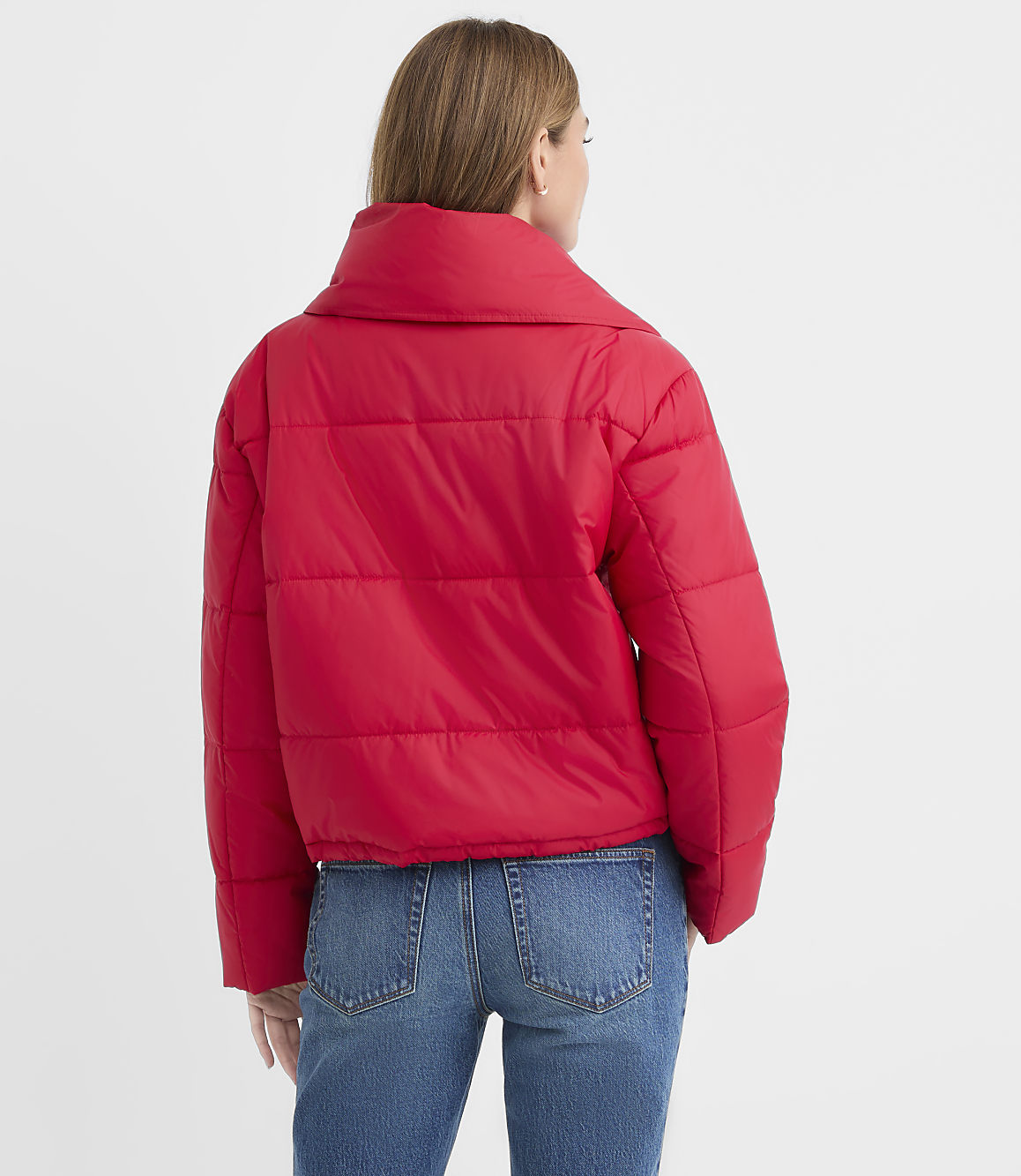 A woman wearing a red puffer jacket and blue jeans stands with her back to the camera against a plain white background.