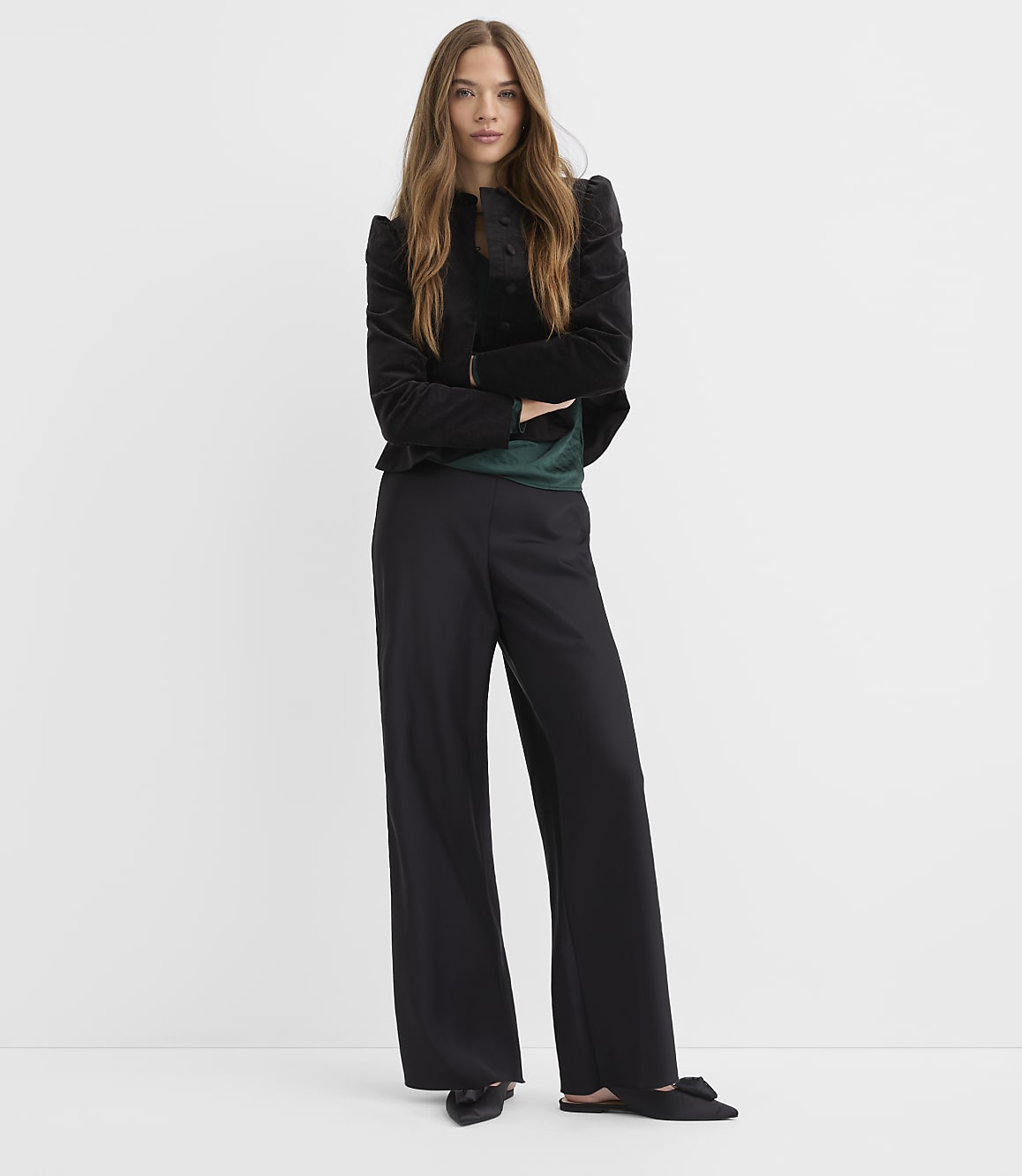 A woman wearing a black velvet jacket, green top, and wide-leg black trousers stands against a plain white background.