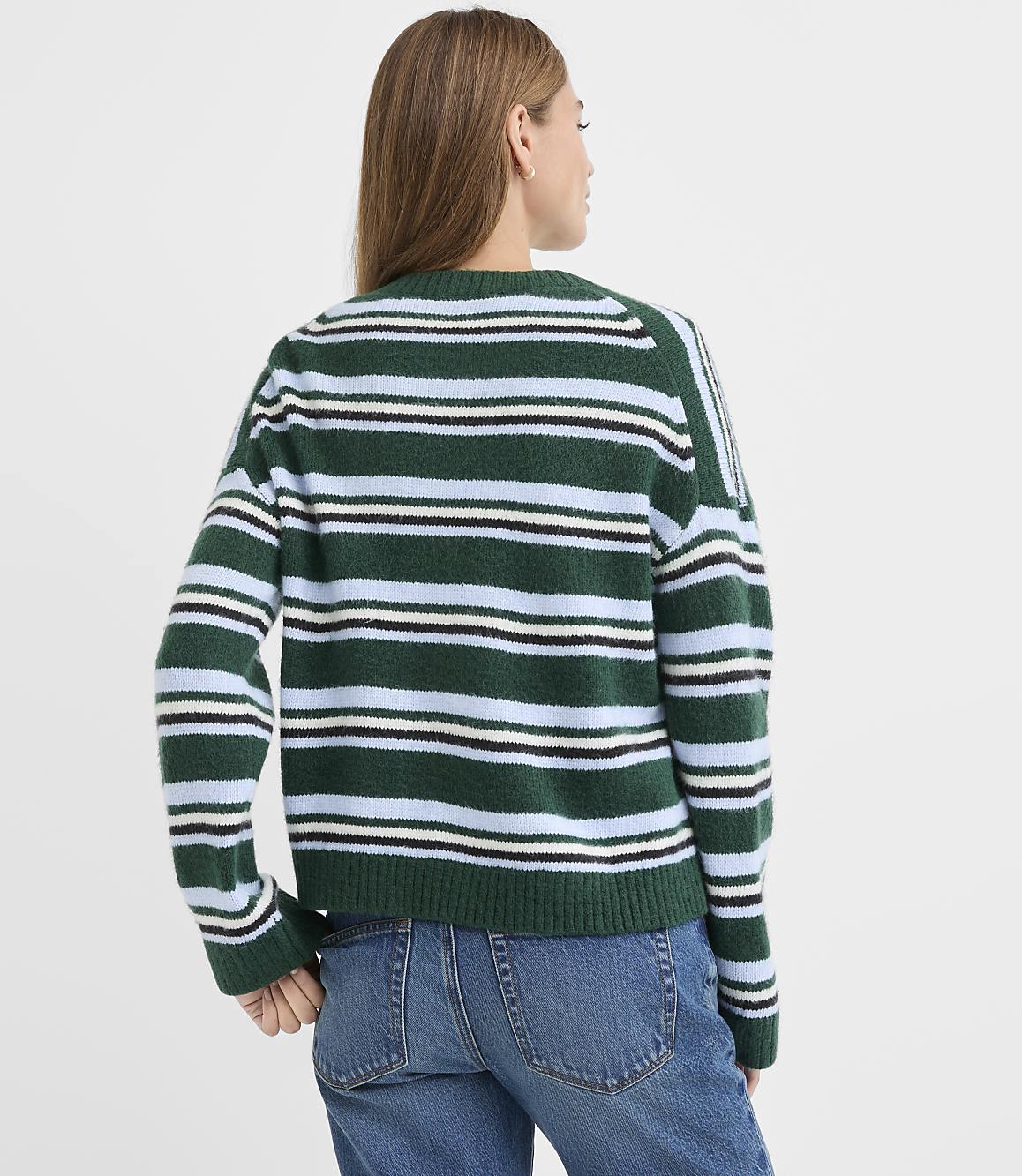 A woman wearing a green, blue, and white striped sweater and blue jeans stands against a plain white background.