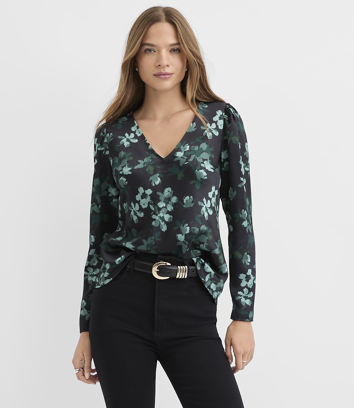 A woman wearing a black floral blouse and black pants stands against a plain white background.