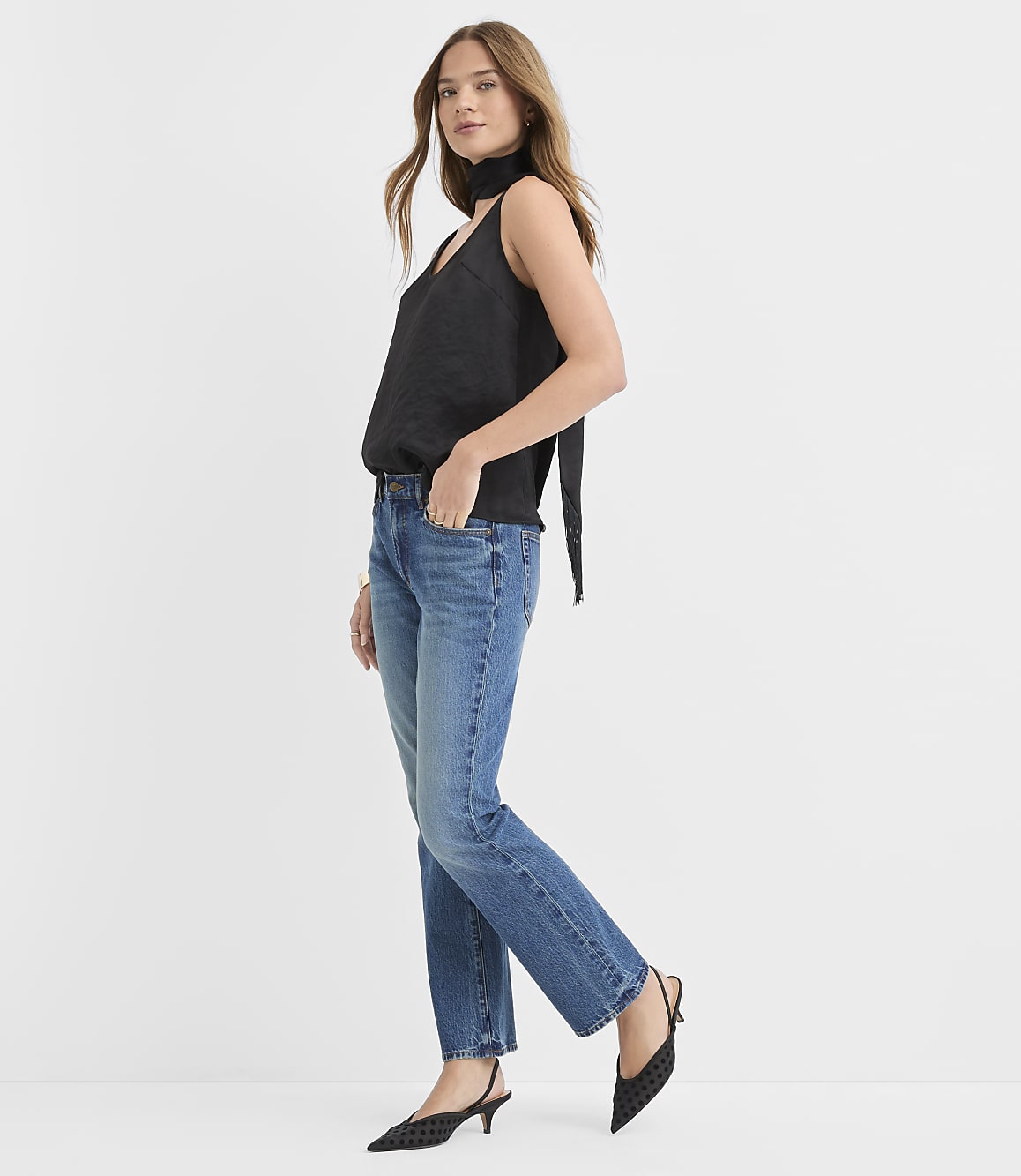 A woman wearing a black sleeveless top, blue jeans, and black pointed heels stands against a plain white background.
