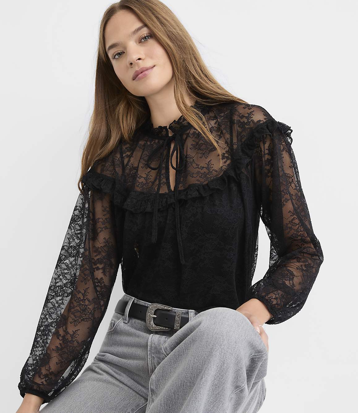 A woman wearing a black lace blouse and grey jeans poses against a plain white background.