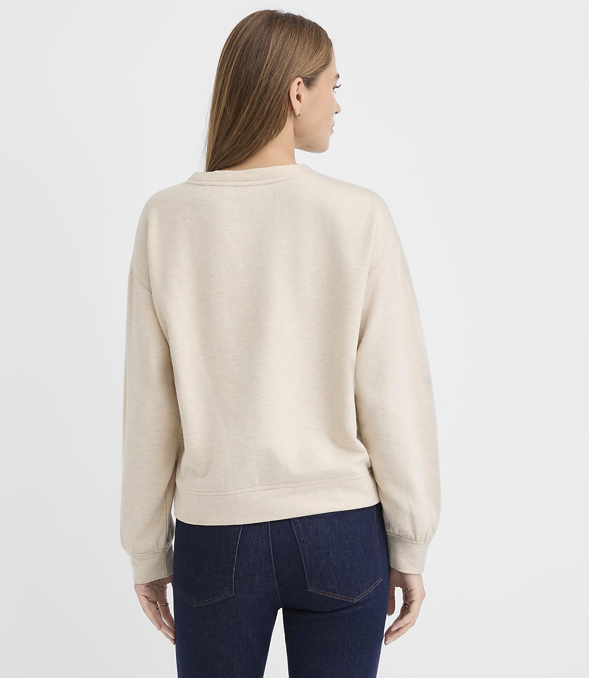 A woman wearing a beige sweatshirt and dark blue jeans stands with her back to the camera against a plain white background.