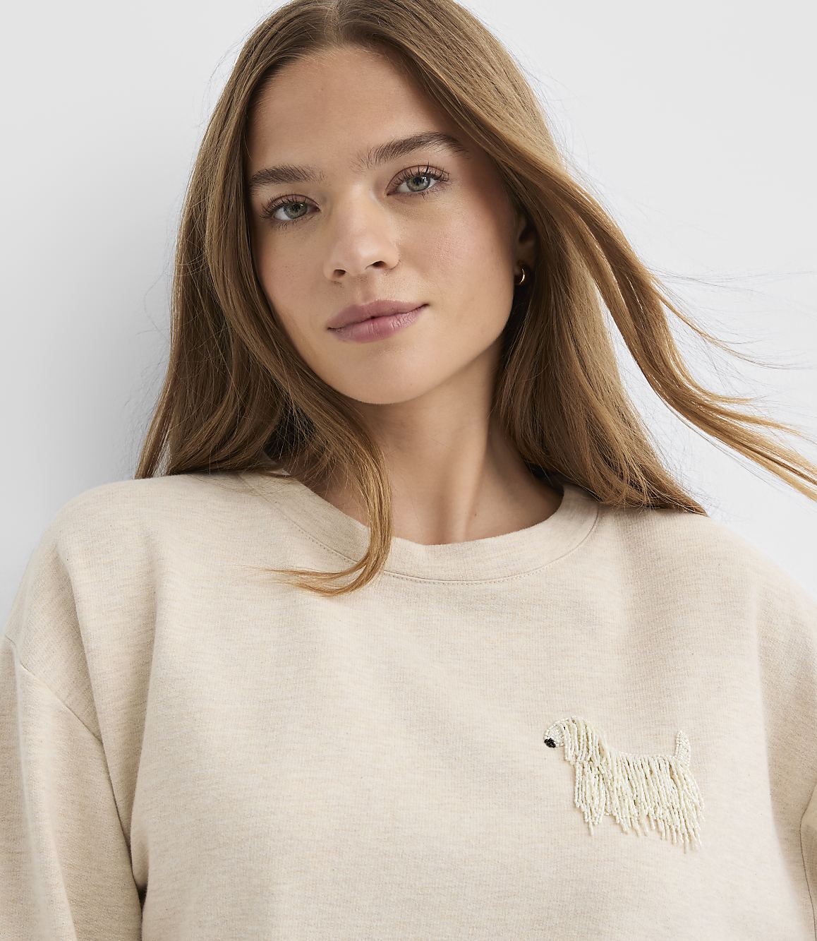 A woman with long brown hair wearing a beige sweatshirt with a small dog embroidery stands against a plain white background.