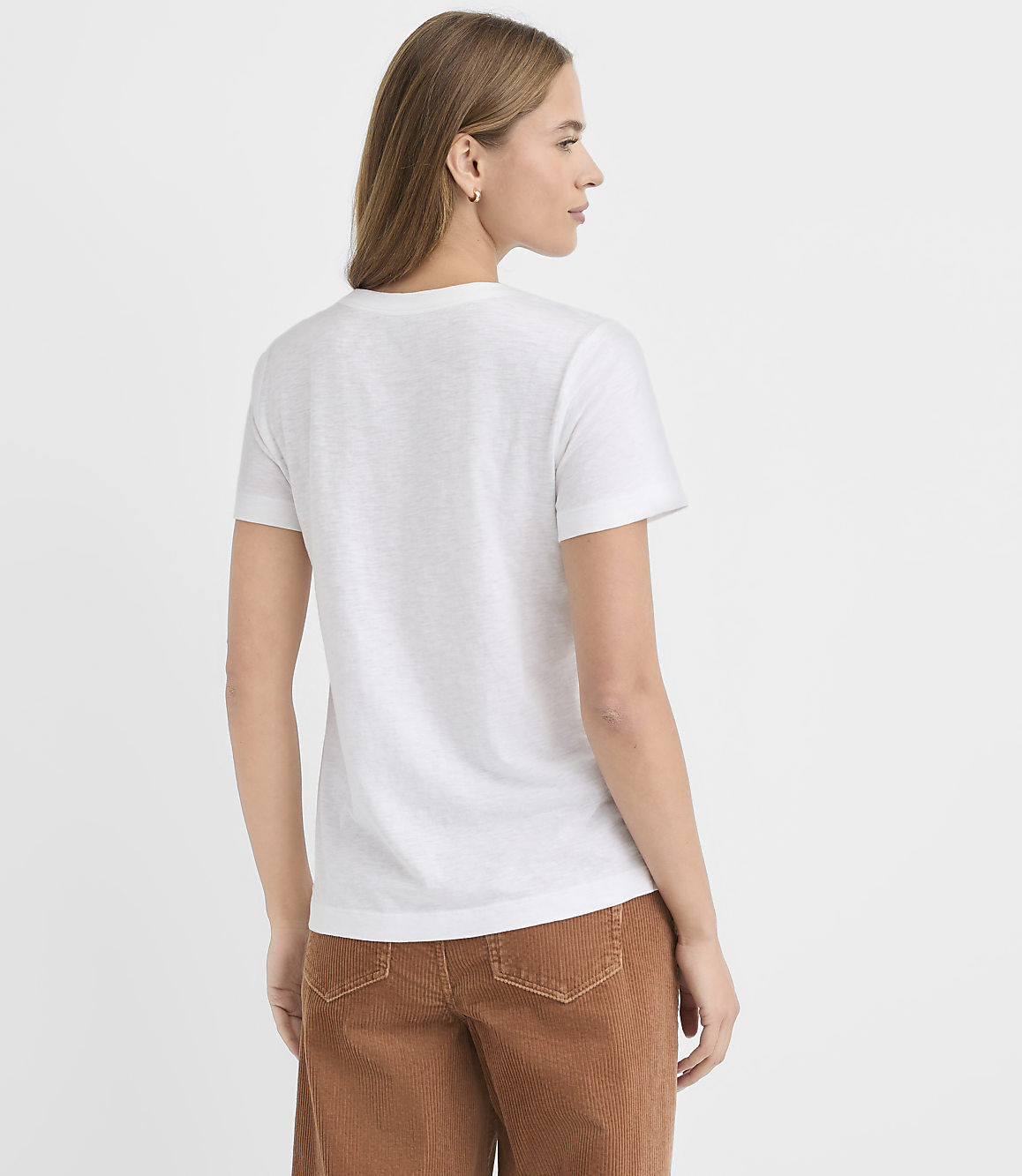 A woman wearing a white t-shirt and brown corduroy pants stands against a plain white background.