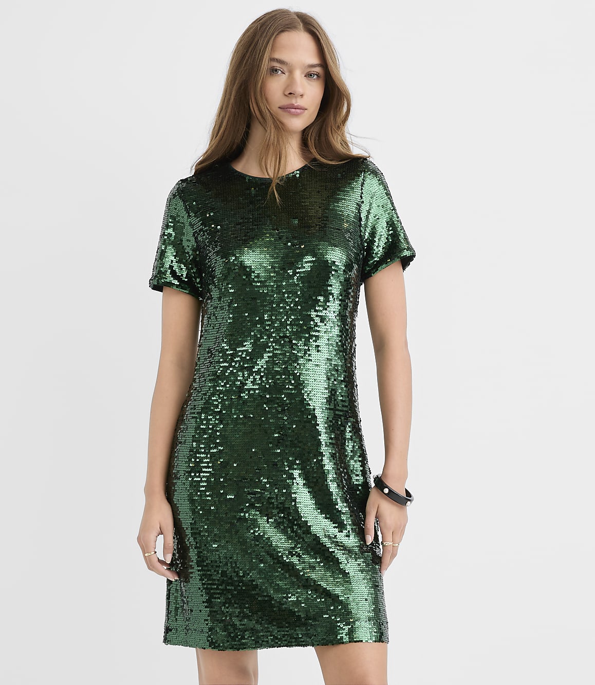 A woman wearing a short-sleeved green sequin dress stands against a plain white background.