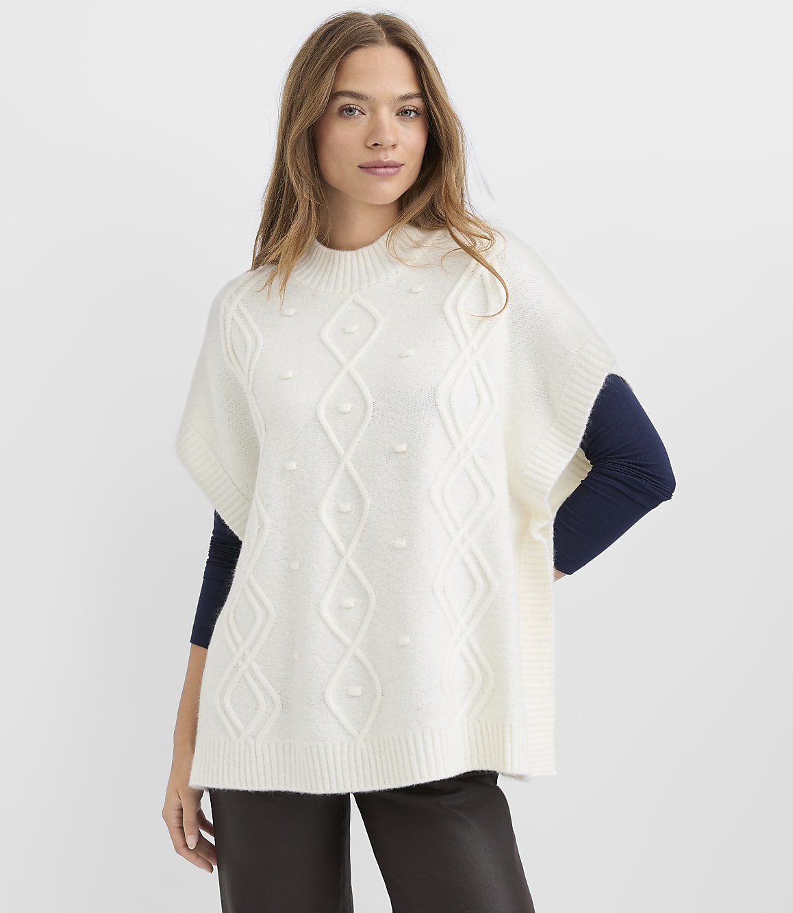 A woman wearing a white cable-knit poncho over a navy blue long-sleeve shirt and dark trousers stands against a plain white background.