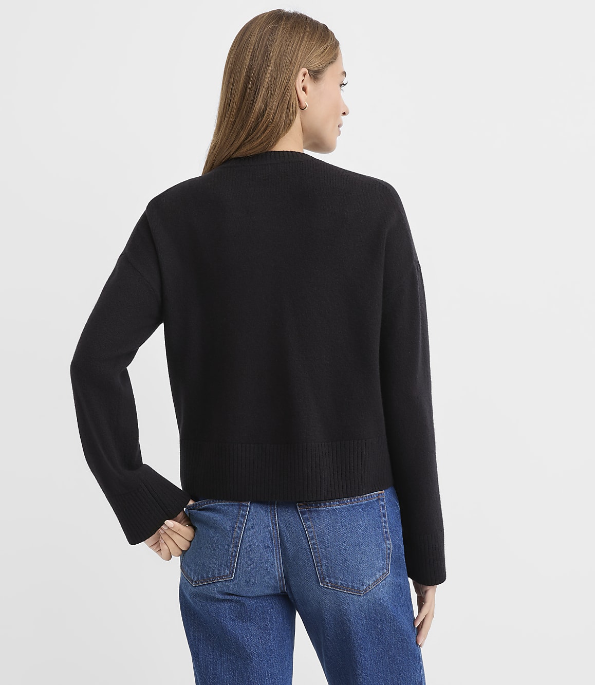 A woman wearing a black sweater and blue jeans stands with her back to the camera against a plain white background.