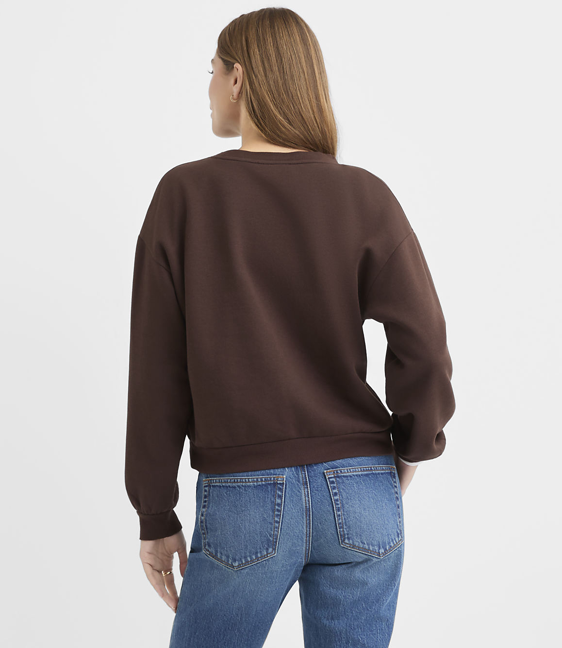 A woman wearing a brown sweatshirt and blue jeans stands with her back to the camera against a plain white background.