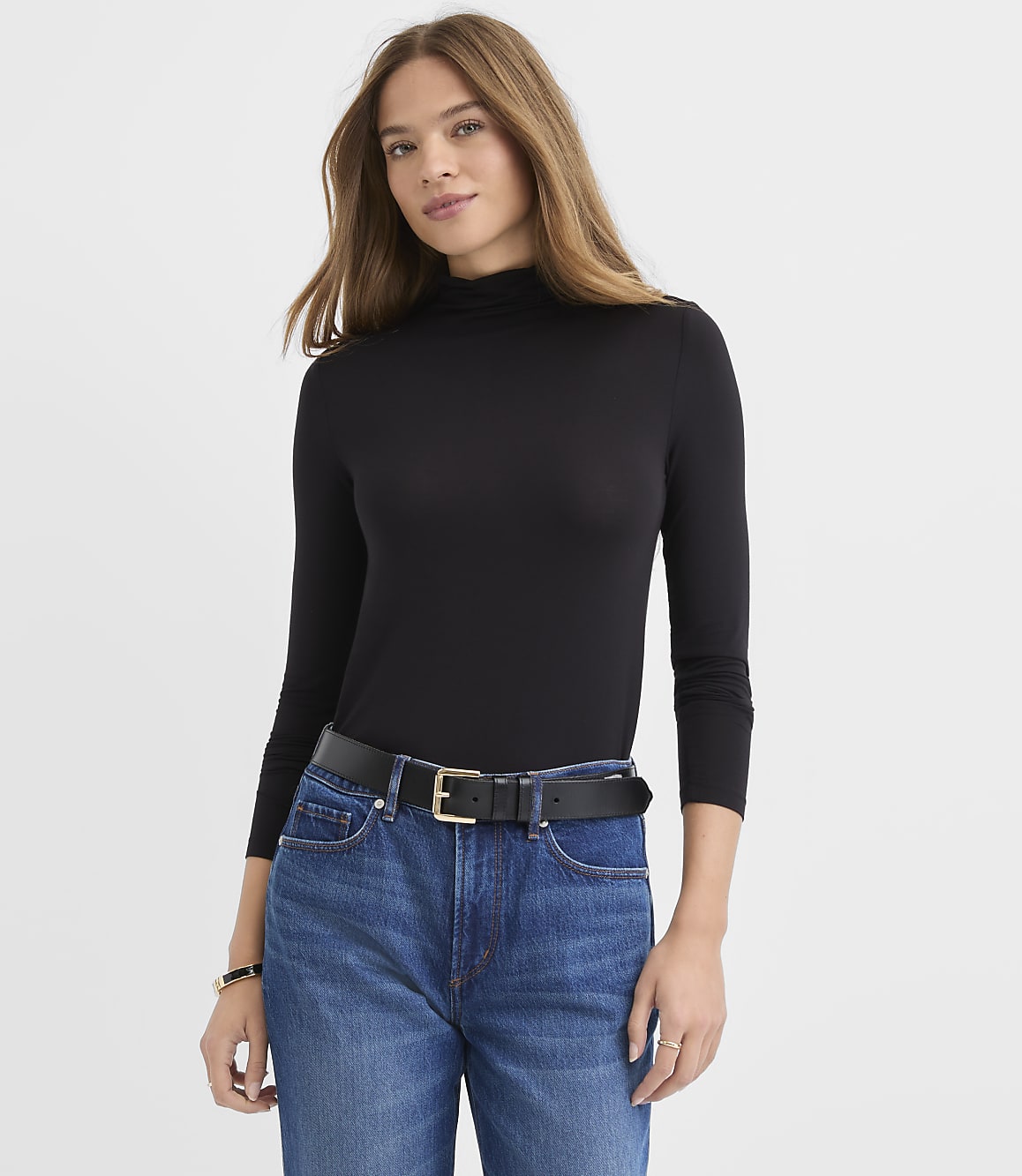 A woman wearing a black turtleneck and blue jeans stands against a plain white background.