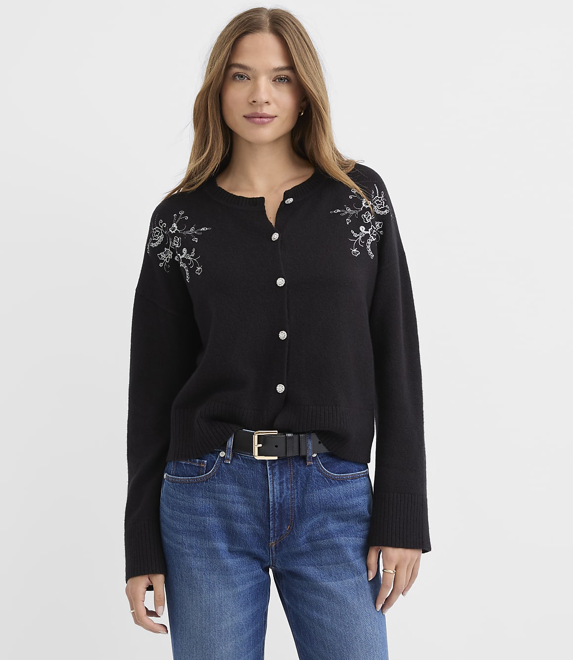 A woman wearing a black embroidered cardigan and blue jeans stands against a plain white background.