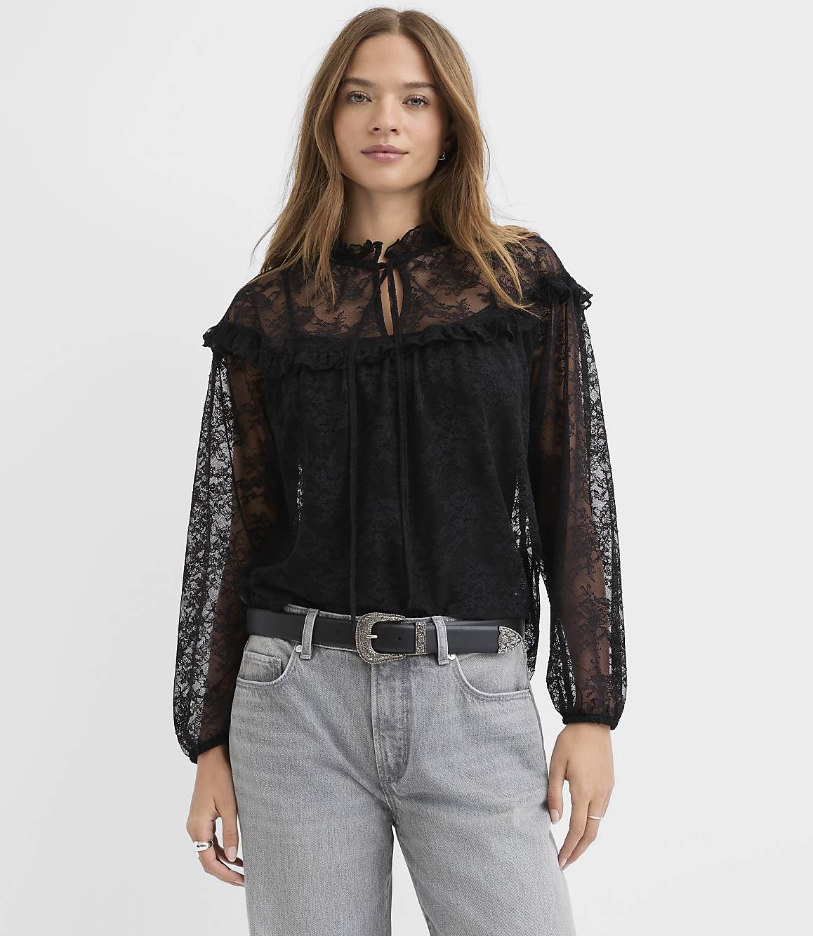 A woman wearing a black lace blouse and grey jeans stands against a plain white background.