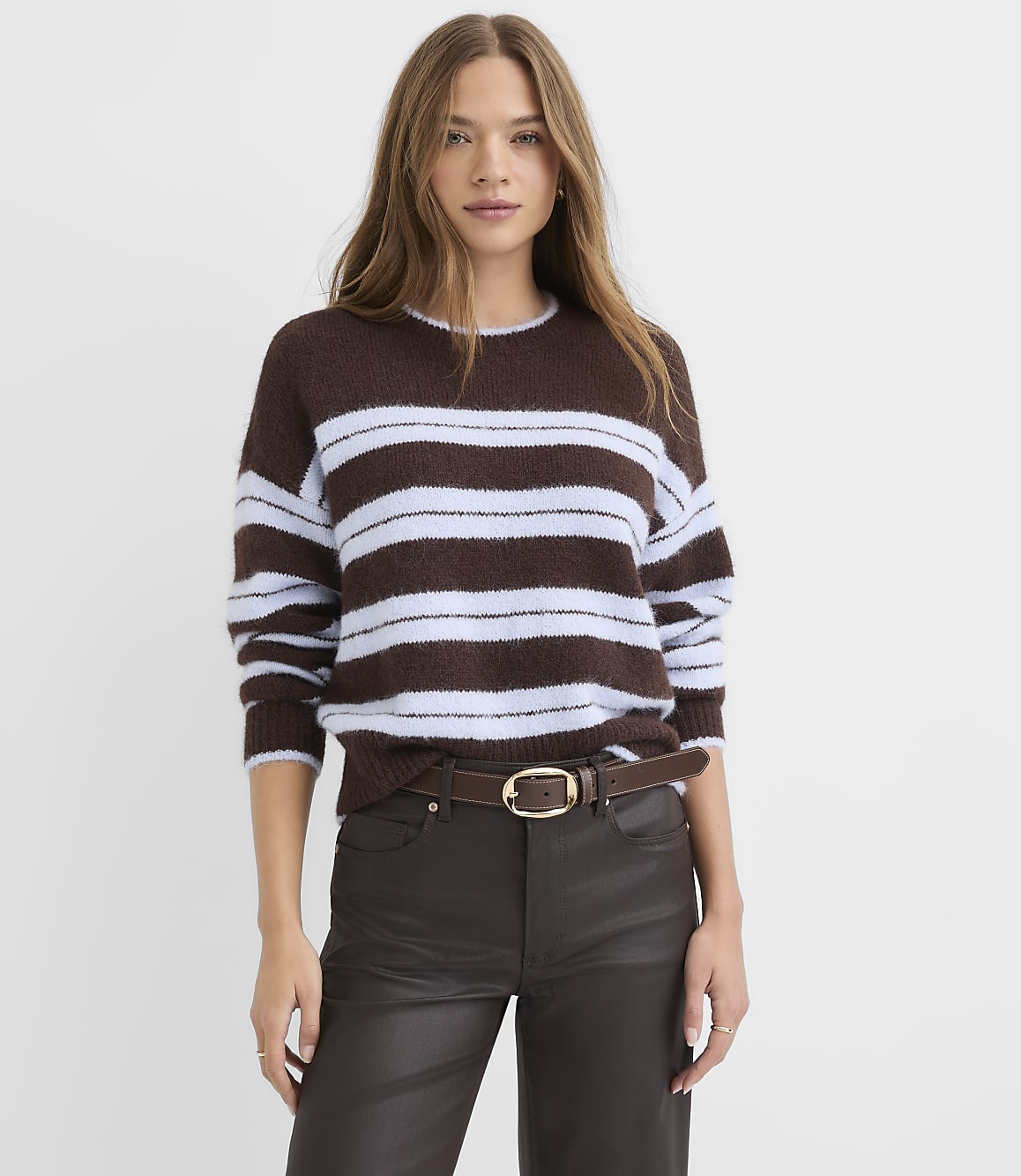 A woman wearing a brown and light blue striped sweater and dark brown leather pants stands against a plain white background.