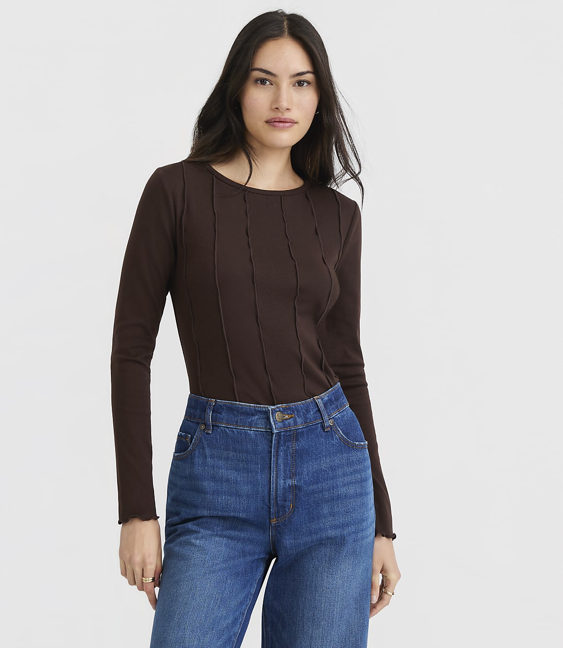 A woman in a brown ribbed top and dark brown pants stands against a plain white background.